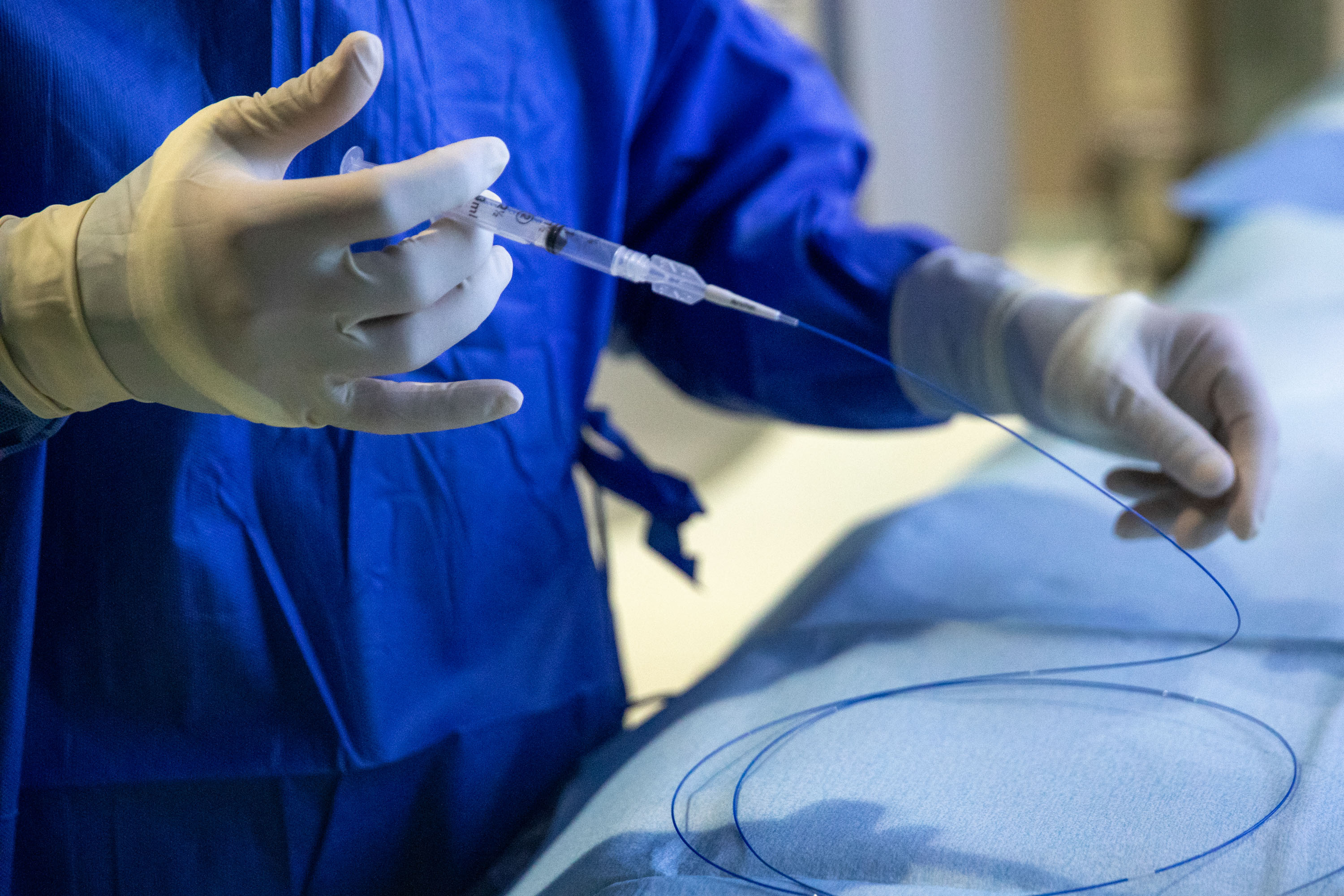  Medical professional in blue scrubs holding catheter and syringe during interventional procedure with patient on examination table.