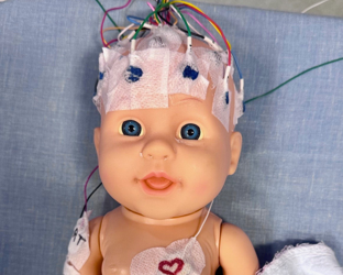 Medical training doll with EEG electrodes and monitoring equipment attached, lying on blue examination surface under blanket.