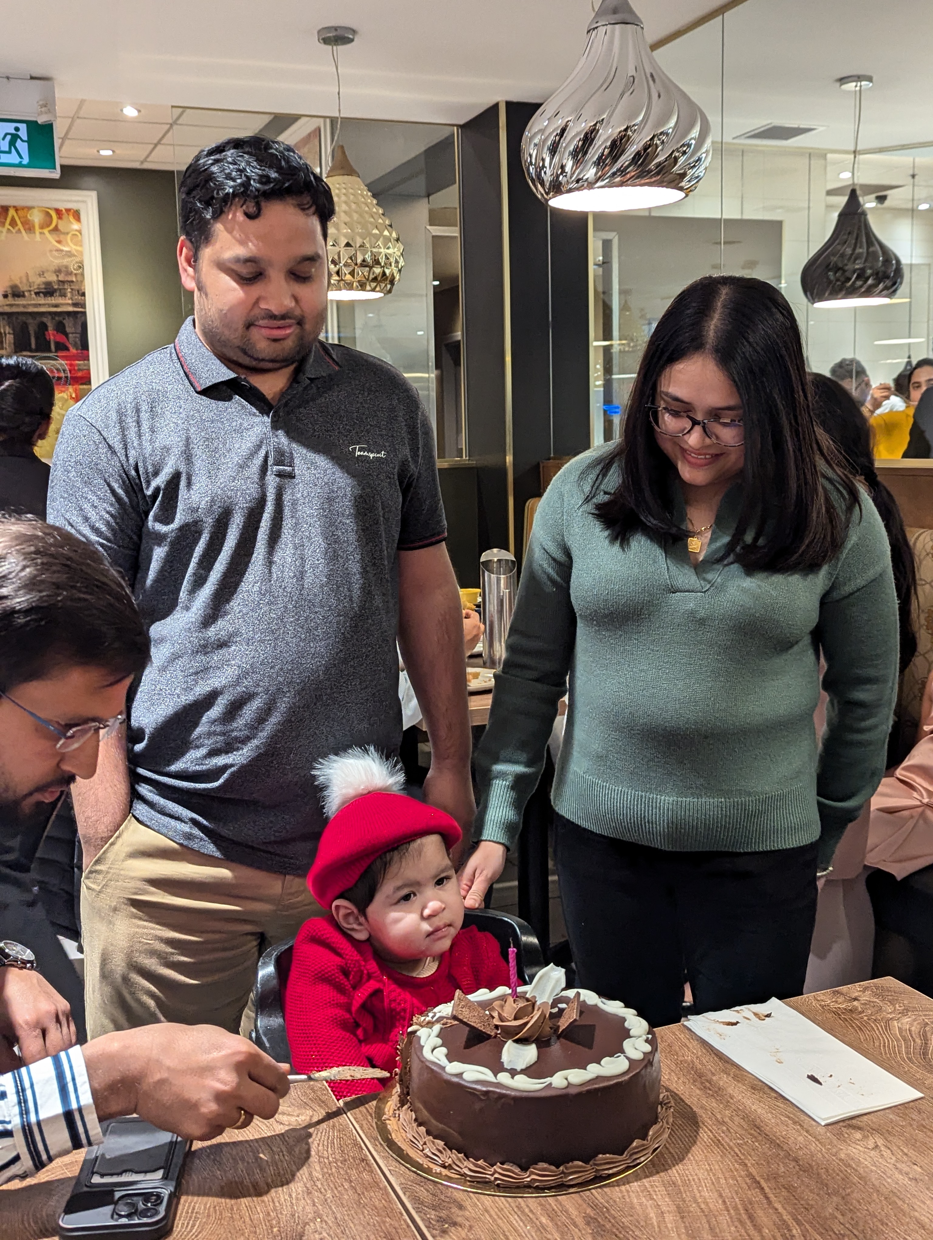 Family celebrating child's first birthday with rose gold balloons, "Happy Birthday" banner, and toddler in party hat surrounded by toys.
