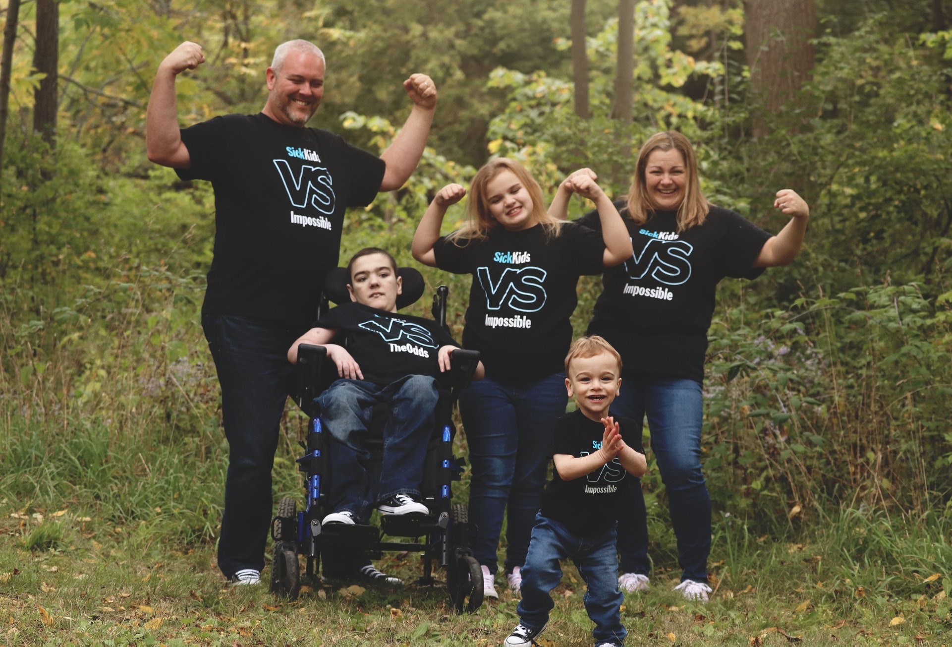 Noah, who is in a wheelchair, and his family wear "SickKids VS" t-shirts, by a green forest.
