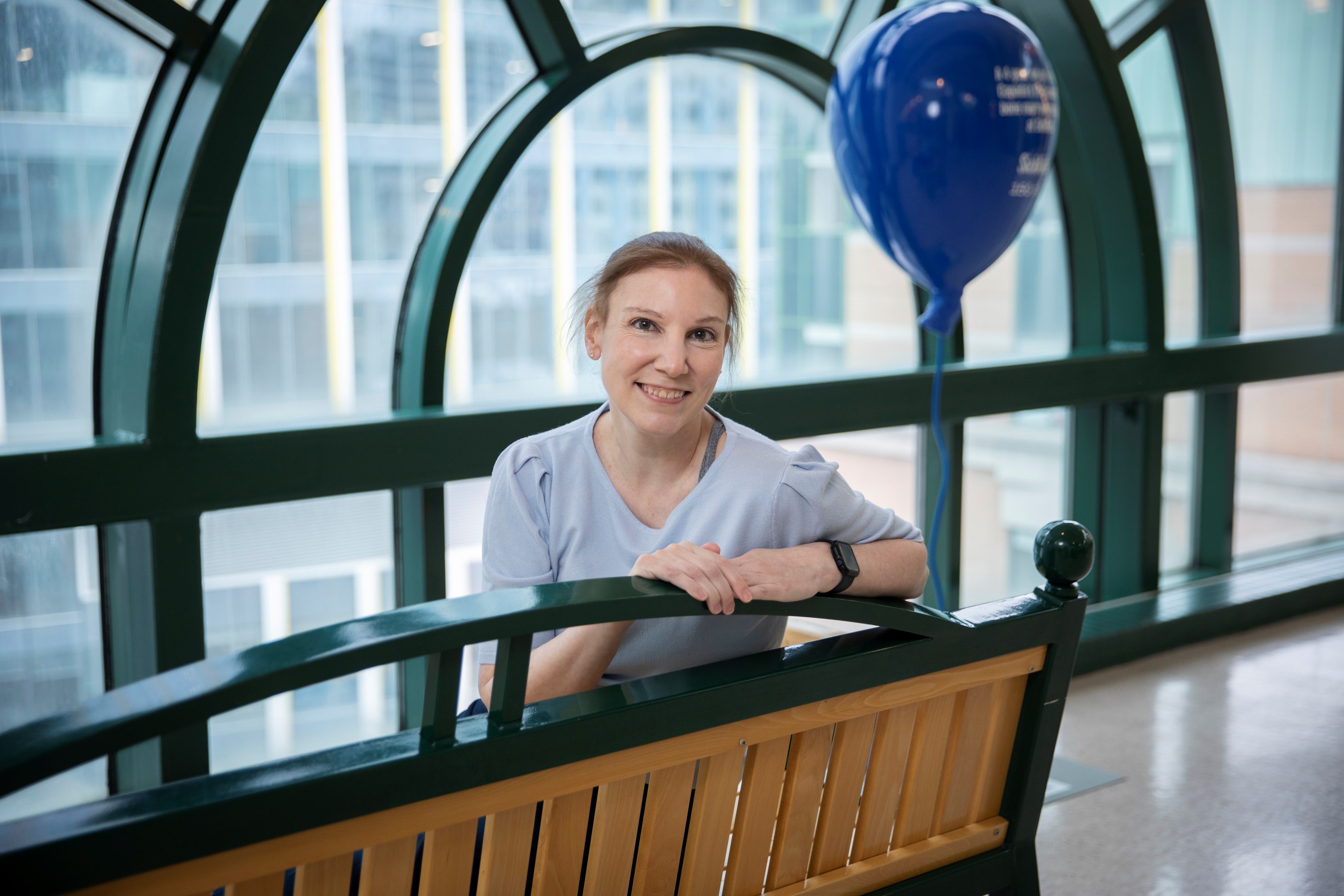 Natalie, now an adult, sits on the same bench. In the background there is a blue balloon sculpture, part of the hospital's 150th birthday celebrations.