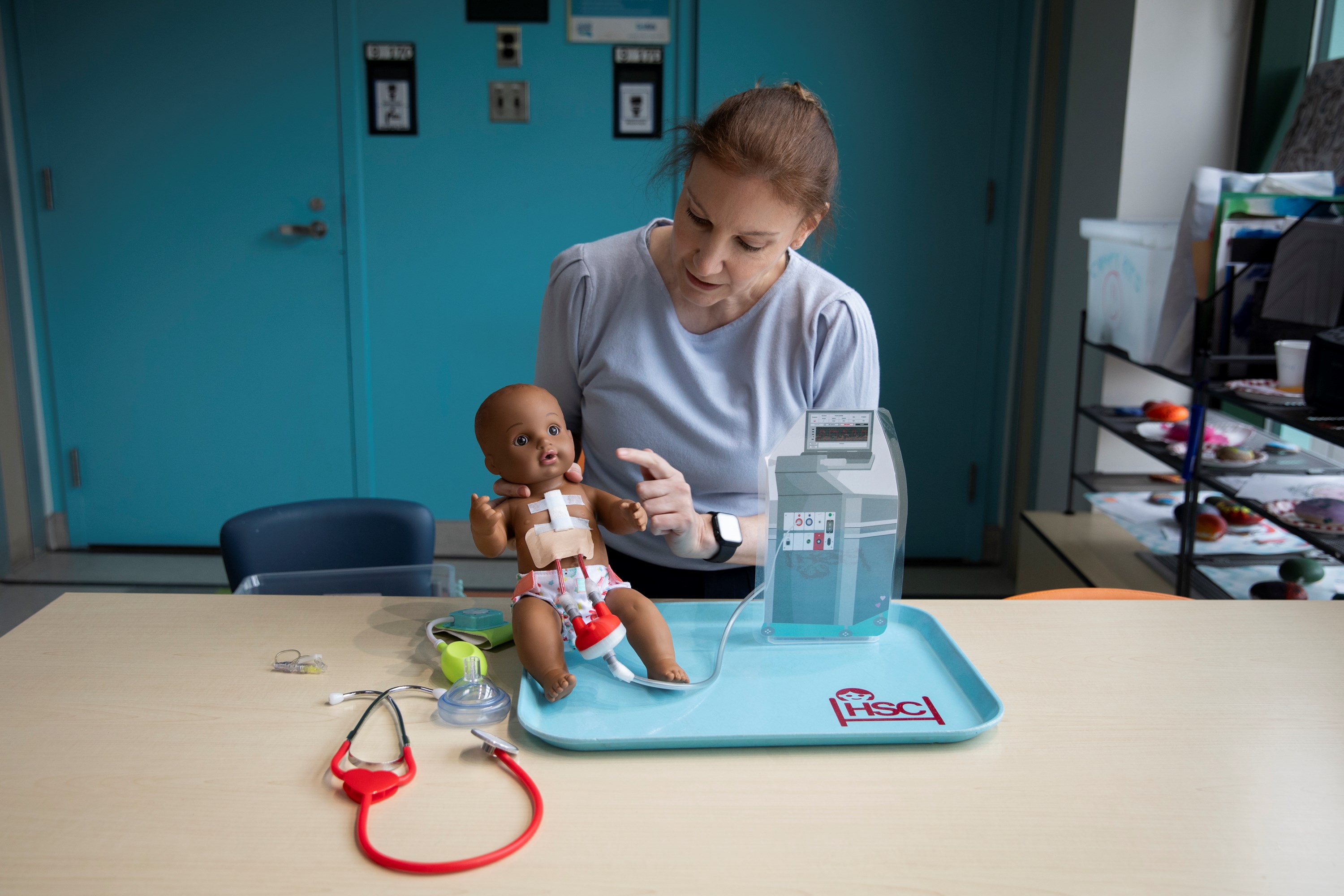 Natalie sits at a table and points at a doll that has a Ventricular Assist Device. There is a stethoscope and other medical equipment on the table.