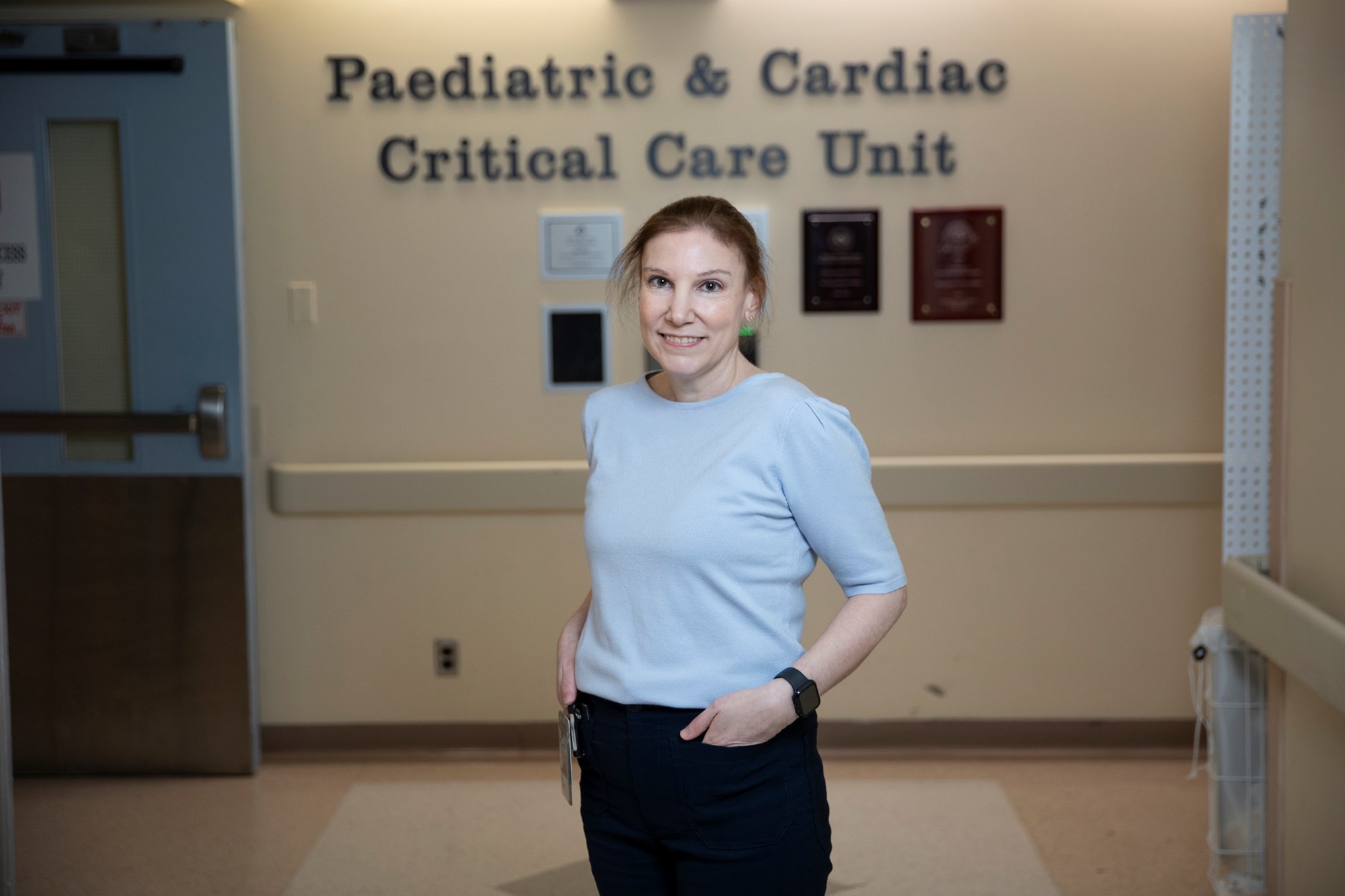 Natalie, in 2025, stands in front of the sign in the unit entrance that reads Paediatric & Cardiac Critical Care Unit.
