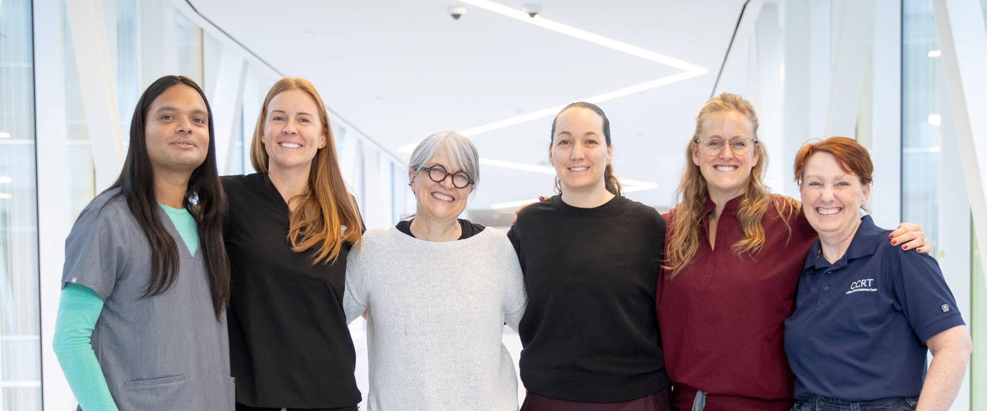 Six healthcare professionals standing together in a bright glass walkway, wearing various medical uniforms.
