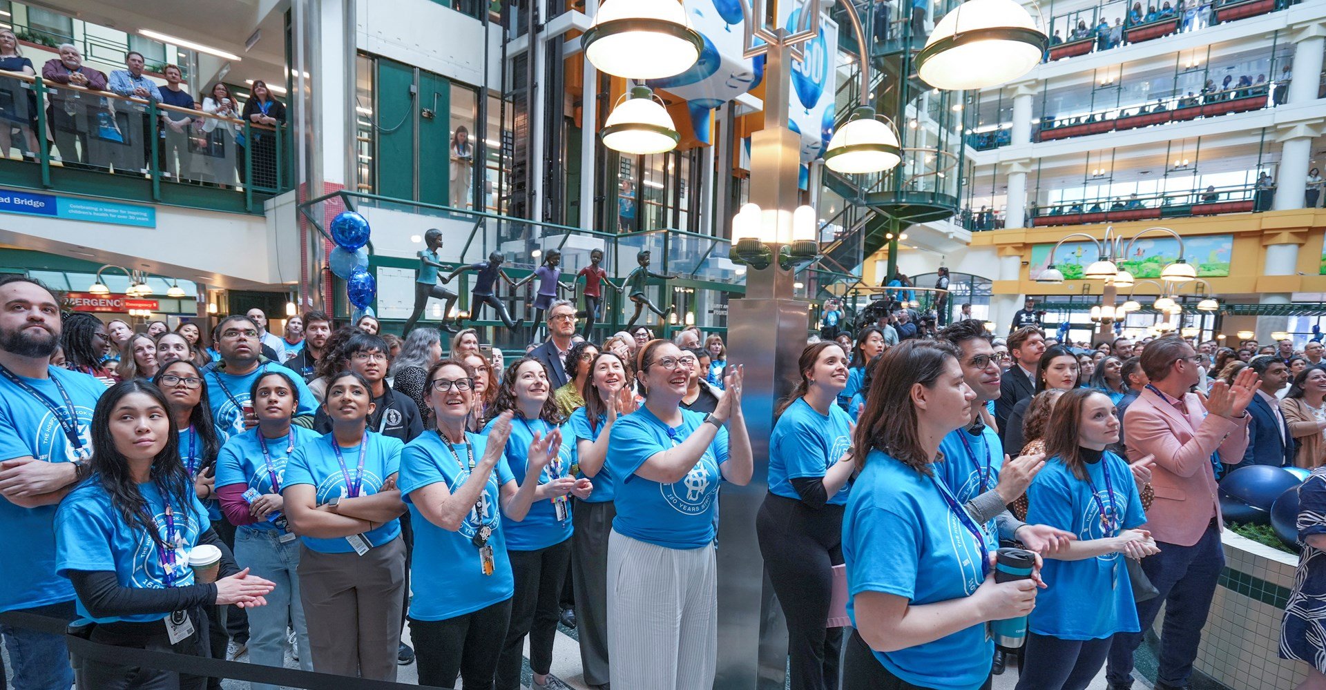 A crowd of people dressed in blue commemorative SickKids 150 t-shirts applauding in the Atrium.