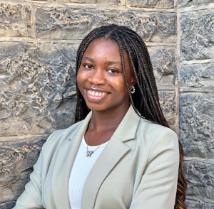 Comfort Oluwagbamila stands in front of a gray brick wall with her arms crossed, wearing a light gray blazer. She is shown from the waist up.