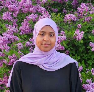 Fatima Ali stands in front of green and light purple foliage with flowers. She is shown from the shoulders up and is wearing a dark dress and light purple hijab.