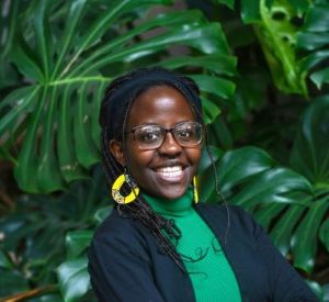 Kimberly Nabukenya stands in front of green foliage with her arms crossed. Se is shown from the waist up.