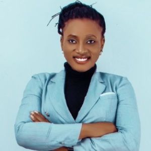 Nayina Mary Bala stands in front of a light teal wall with her arms crossed. She is wearing a light teal blazer and is shown from the waist up.
