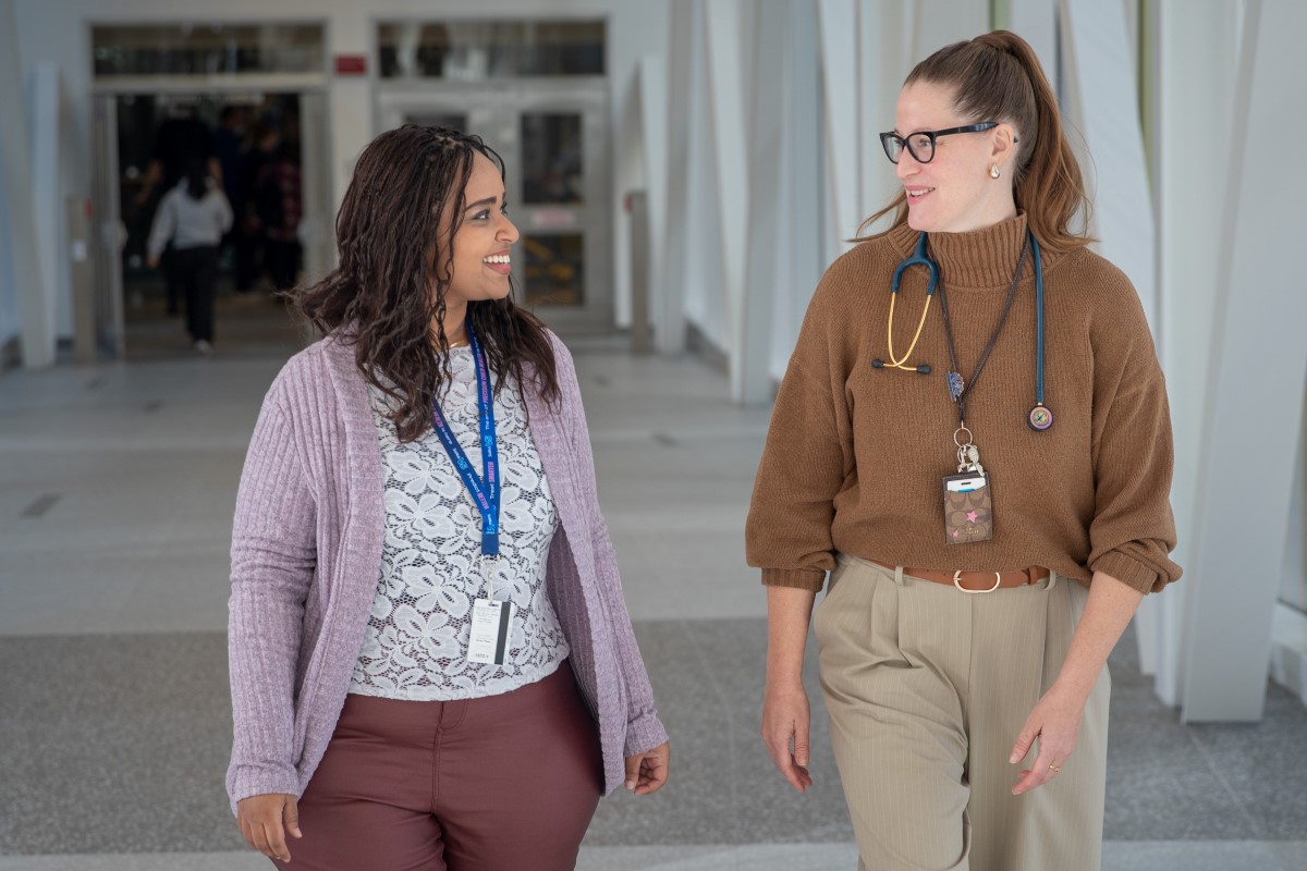 Two healthcare workers wearing ID badges walk side by side through a bright hospital hallway, talking as they move toward an entrance.