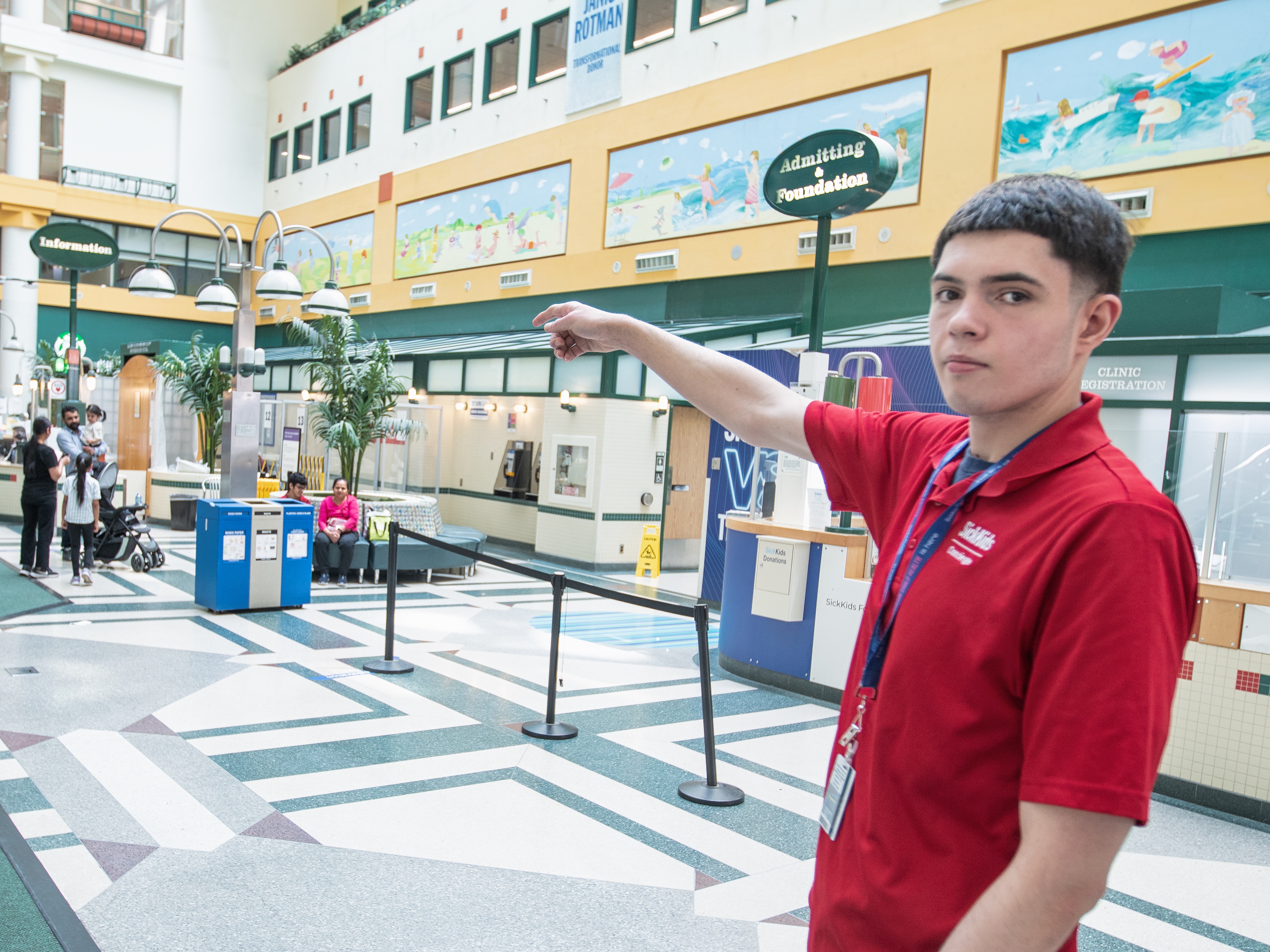 A man in a red shirt points across a large hospital atrium with seating, signage, and people in the background.
