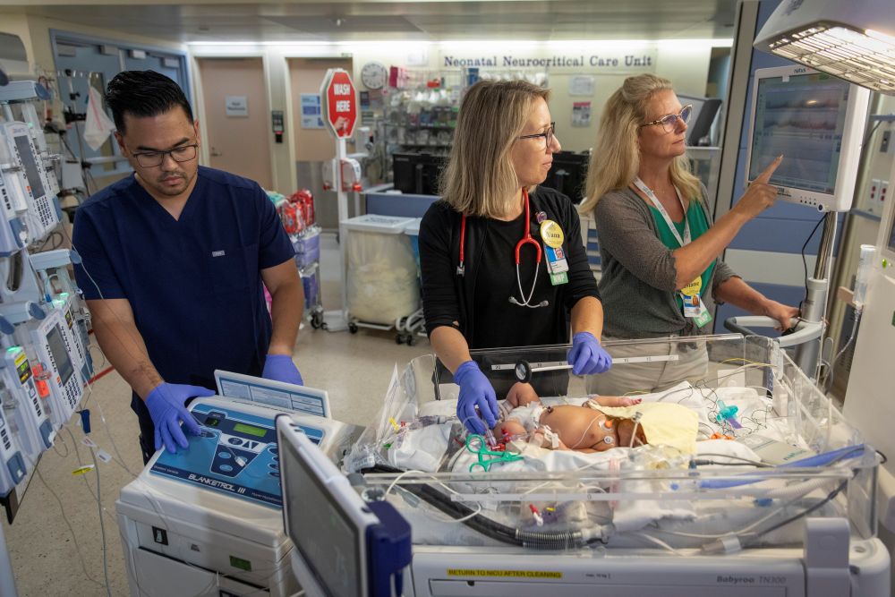 Three nurses surround an infant in a NICU bed as they work with or look at medical equipment.