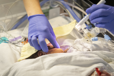 Two hands with medical gloves are shown touching and assessing an infant patient in the NICU.
