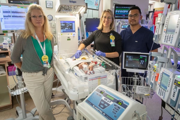 Three nurses pose for the camera and stand around an infant patient sleeping in a NICU bed.