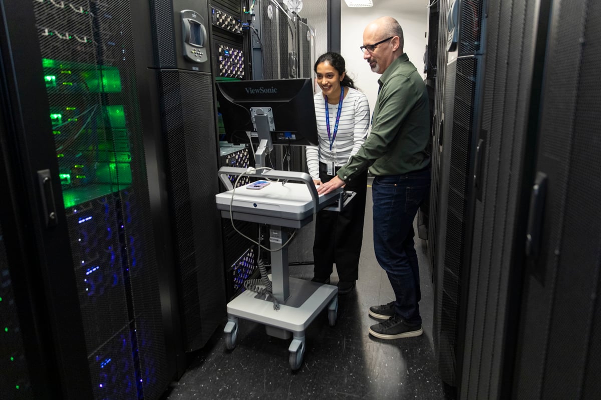 Two technicians stand in a data centre aisle, working at a mobile workstation surrounded by tall server racks with glowing indicator lights