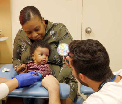 Woman holds baby. Man kneels facing the baby holding a toy.