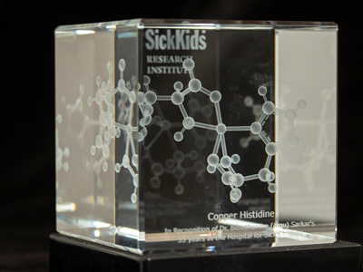 A molecular model of a copper histidine molecule encased in a glass cube. The text engraved on the cube reads, "Copper Histidine. In recognition of Dr. Bibudhendra (Amu) Sarkar's 55 years at The Hospital for Sick Children