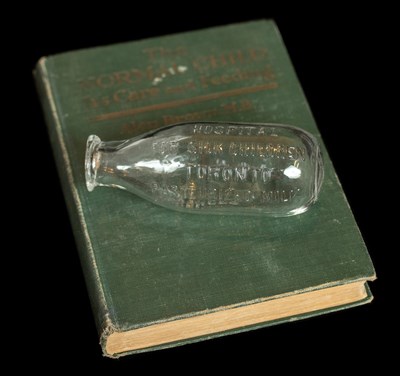A glass milk bottle with a curved bottom. The lettering on the glass reads, "Hospital for Sick Children Toronto Pasteurized Milk" . The bottle is sitting on its side on top of a worn green book.