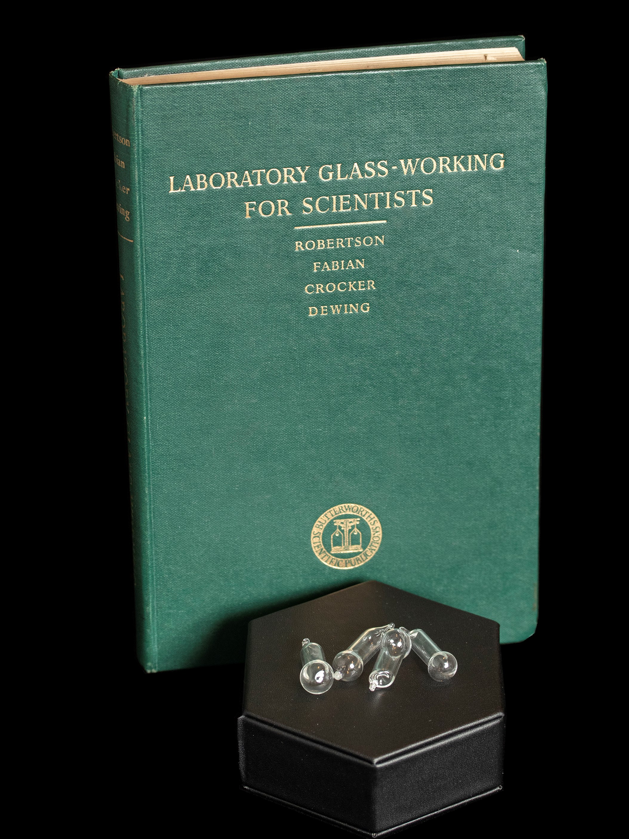 A green book titled "Laboratory Glass-Working for Scientists" standing upright behind a black hexagonal case. On top of the case are four glass test tube stoppers.