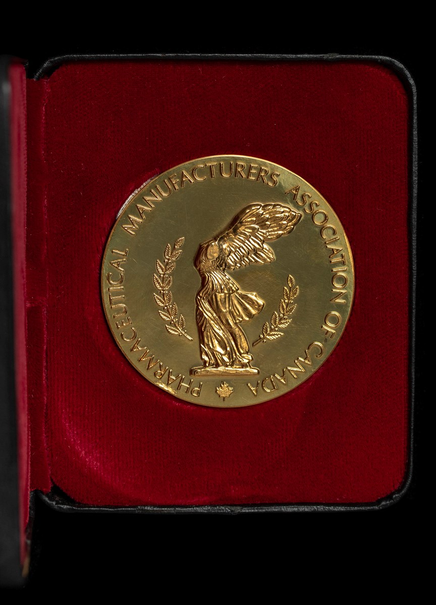 A gold medal of a headless angel with its wings spread. The figure is surrounded by two laurels. The engraved text reads, "Pharmaceutical Manufacturers Association of Canada". 