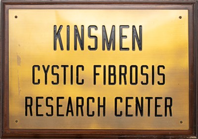 A wooden plaque with metal plating on which "Kinsmen Cystic Fibrosis Research Center" is engraved.