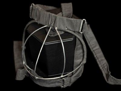 A mask standing upright. The mask is made of cloth straps to secure around a child's head and a metal wire piece that goes over the bottom half of the child's face that prevents them from eating lead paint.