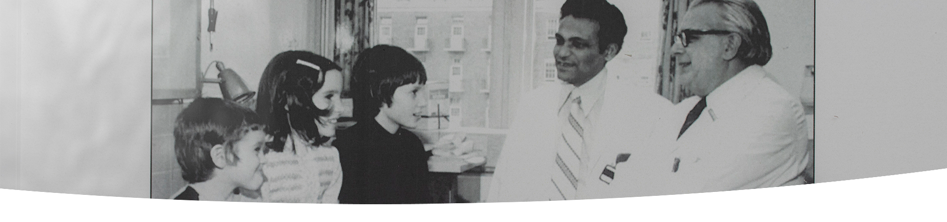 Black and white photo of Dr. Bibudhendra (Amu) Sarkar and Dr. Andrew Sass-Kortsak speaking with three children with Wilson disease