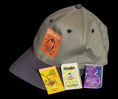 A baseball cap with the Sprinkles logo (an orange square with a smiling baby in the centre). Three packets of Sprinkles sit on top of the baseball cap.
