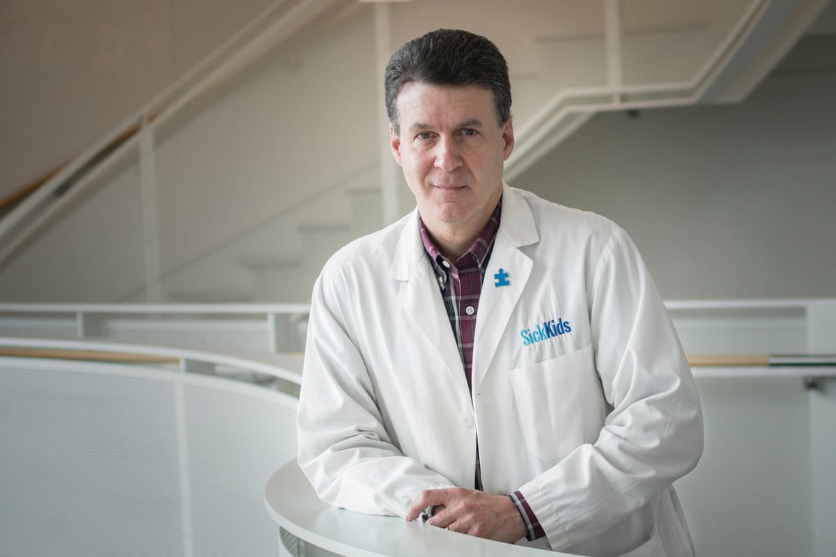 Dr. Stephen Scherer in a white SickKids lab coat standing with arms crossed in a laboratory, with researchers and scientific equipment visible in the background